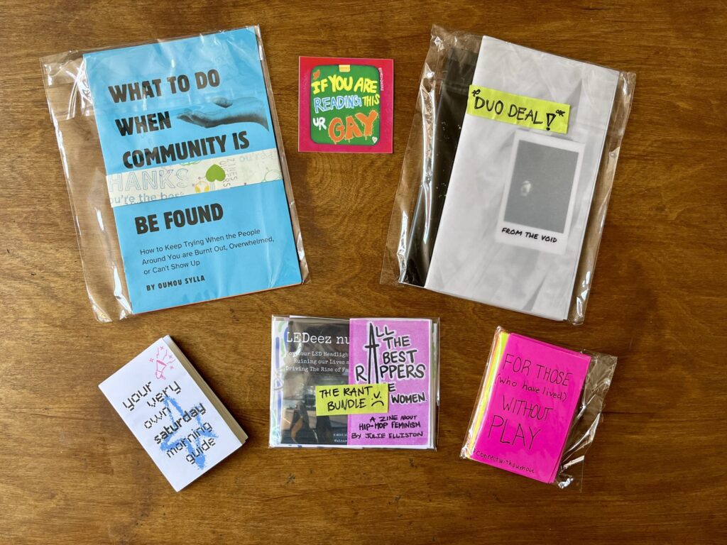 In Cellophanhüllen verpackte Zines von Zines4Queers liegen auf einem Holztisch. Die vier Bundles "Community", "The Rant Bundle", "Duo Deal" und "For Those Who Have Lived..." sind zu sehen sowie ein Mini-Zine "Your very own saturday morning guide".
