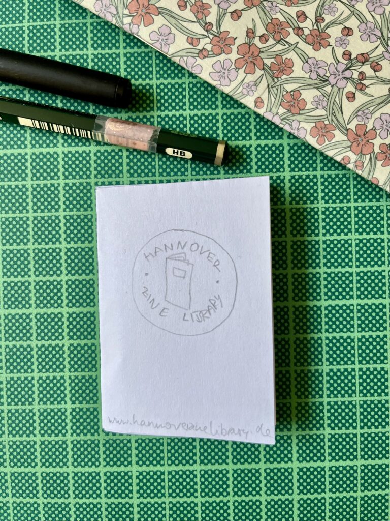 Auf einer grünen Schneidematte liegt ein Prototyp eines Zines über die HZL. Es ist das mit Bleistift gezeichnete Cover zu sehen: das kreisförmige HZL-Logo und am unteren Rand des Zines steht die URL. Als Größenindes wurde ein Bleistift und ein schwarzer Stift ins Bild gelegt.