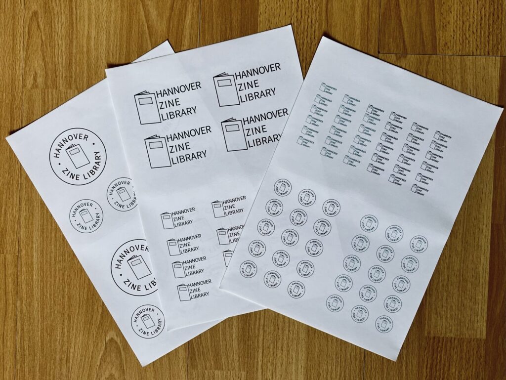Auf einem dunklen Holz liegen drei Stickerbögen mit den zwei unterschiedlichen HZL-Logo-Designs in verschiedenen Größen.