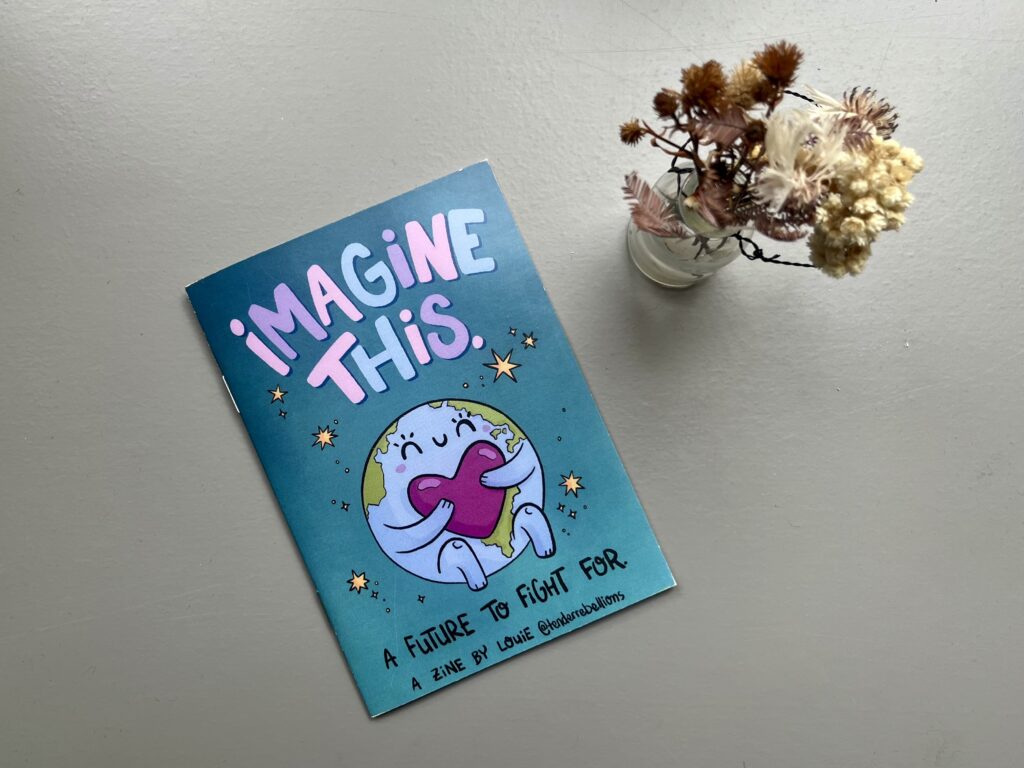 Auf einer grauen, glatten Oberfläche liegt das Zine „Imagine This. A Future to Fight For.“ von Louie Läuger. Daneben steht ein kleiner Trockenblumenstrauß. Das Motiv wurde aus einer Draufsicht fotografiert.